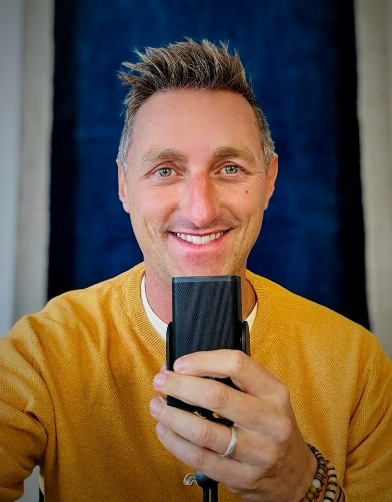 Chris Spieler hält ein Mikrofon in den Händen und lächelt – ein Moment liebevoller Präsenz beim Einsprechen individueller Meditationen.